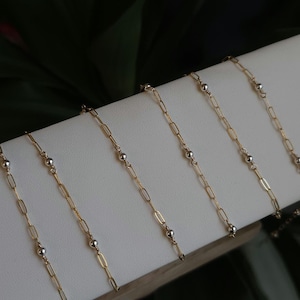 May include: Six gold chains with silver beads, displayed on a white surface. The chains are of varying lengths and are arranged in a staggered pattern.
