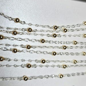 Puede incluir: Varias hebras de cadena de plata con cuentas doradas. Las cadenas est&aacute;n superpuestas y entrelazadas, creando un dise&ntilde;o delicado e intrincado.