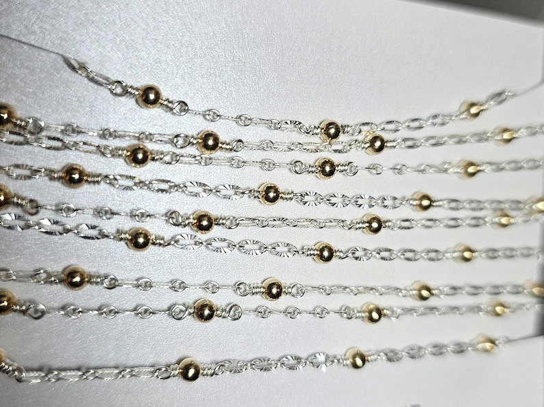Puede incluir: Varias hebras de cadena de plata con cuentas doradas. Las cadenas est&aacute;n superpuestas y entrelazadas, creando un dise&ntilde;o delicado e intrincado.