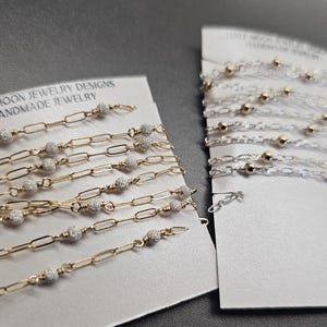 Puede incluir: Dos tarjetas con cadenas doradas y plateadas con peque&ntilde;as cuentas. La tarjeta de la izquierda dice "Half Moon Jewelry Designs Handmade Jewelry". La tarjeta de la derecha dice "Half Moon Jewelry Designs Handmade Jewelry".