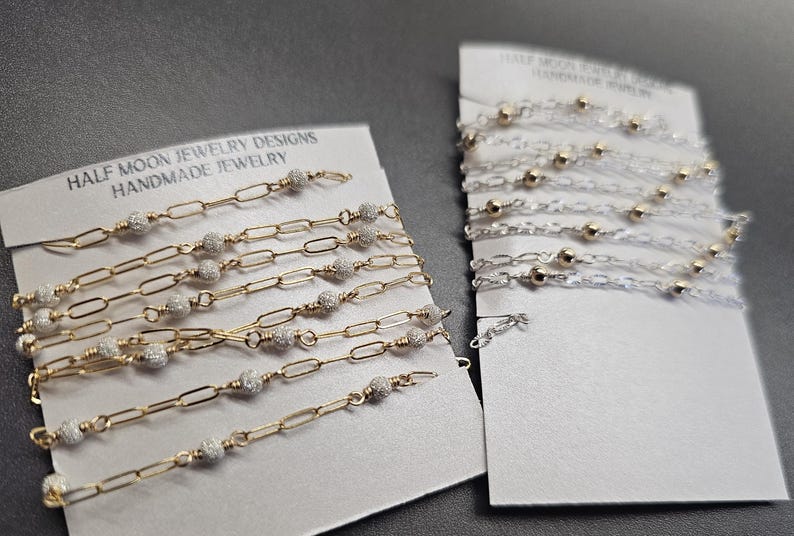 Puede incluir: Dos tarjetas con cadenas doradas y plateadas con peque&ntilde;as cuentas. La tarjeta de la izquierda dice "Half Moon Jewelry Designs Handmade Jewelry". La tarjeta de la derecha dice "Half Moon Jewelry Designs Handmade Jewelry".