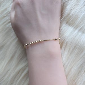 May include: A delicate gold bracelet with small, evenly spaced beads and a slender bar. The bracelet is secured with a small clasp. The jewelry is displayed on a wrist against a soft, light-coloured background.