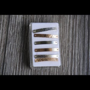 以下が含まれることがあります： 透明なプラスチックケースに入った長方形の金属バーのコレクション。バーはシルバー、ゴールド、ローズゴールドなど、さまざまな色があります。ハンマーで叩いたような質感のバーもあれば、滑らかなバーもあります。各バーには小さな穴があります。