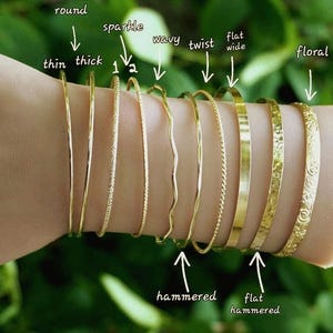 May include: A selection of gold-coloured bangles in diverse styles, including thin, thick, sparkle, wavy, twist, flat, wide, and floral designs. The bangles are shown on a wrist against a blurred green backdrop.