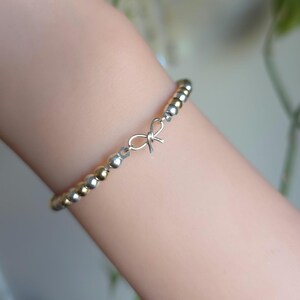May include: A delicate bracelet featuring small silver and gold beads. A small silver bow charm is centered on the bracelet. The bracelet is worn on a wrist.