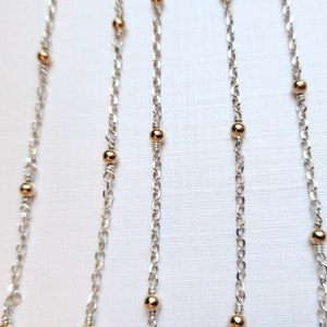 May include: Four silver chains with gold beads. The chains are hanging straight down against a white background.
