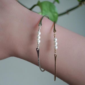 May include: Two delicate bracelets. One is silver-toned with a chain and a pearl accent. The other is gold-toned with a pearl accent. Both bracelets have a textured, elongated charm. The bracelets are on a person's arm.