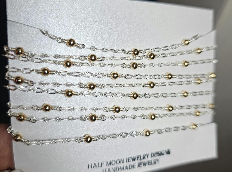Puede incluir: Primer plano de varias cadenas plateadas con cuentas doradas. Las cadenas est&aacute;n superpuestas y se muestran sobre un fondo blanco. El texto "HALF MOON JEWELRY DESIGNS HANDMADE JEWELRY" est&aacute; impreso en la parte inferior de la imagen.