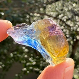 Może przedstawiać: Wielokolorowy, półprzezroczysty kawałek szkła o nieregularnym kształcie. Szkło ma gradient kolorów, w tym niebieski, żółty i fioletowy. Obiekt trzymany jest między dwoma palcami, z rozmytym zielonym tłem.