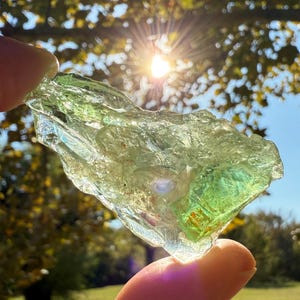 May include: A translucent, irregularly shaped, green crystal is held up to the sunlight. The crystal has a rough, textured surface and varying shades of green. The background features a bright sun, trees, and a grassy area.