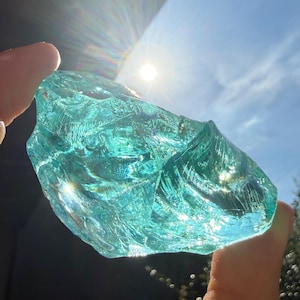 Könnte beinhalten: Ein großes, unregelmäßig geformtes Stück durchscheinendes, türkis gefärbtes Glas wird gegen die Sonne gehalten. Das Licht scheint durch das Glas und erzeugt einen hellen, funkelnden Effekt. Der Hintergrund ist ein klarer blauer Himmel.