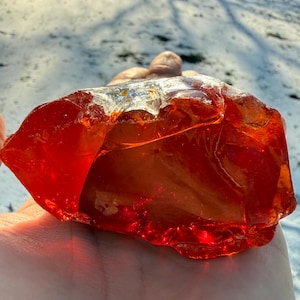 May include: A large, translucent, orange-red glass rock with a rough, uneven surface. The light shines through the glass, creating a glowing effect. The rock is held in a hand, with a snowy background.