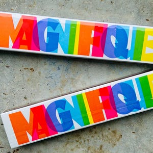 Könnte beinhalten: Zwei rechteckige Streichholzschachteln mit dem Wort "MAGIFIQUE" in großen, farbenfrohen Buchstaben. Die Buchstaben sind in einem Regenbogen von Farben gehalten, mit Orange, Rot, Rosa, Blau, Grün und Gelb. Eine Schachtel ist geöffnet.