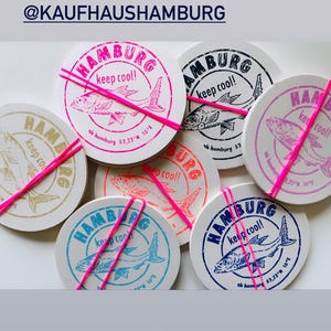 Pode incluir: Uma coleção de bases de copo redondas com uma ilustração de peixe e a palavra "Hamburg" em várias cores, incluindo rosa, azul e laranja. Cada base de copo é presa com um elástico rosa brilhante. O texto "keep cool!" também é visível.