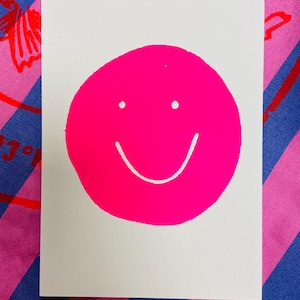 K&ouml;nnte beinhalten: Eine wei&szlig;e rechteckige Karte mit einem gro&szlig;en, leuchtend pinkfarbenen Smiley-Gesicht. Das Smiley-Gesicht hat zwei kleine wei&szlig;e Punkte f&uuml;r die Augen und eine gebogene wei&szlig;e Linie f&uuml;r den Mund. Die Karte befindet sich vor einem gemusterten Hintergrund.