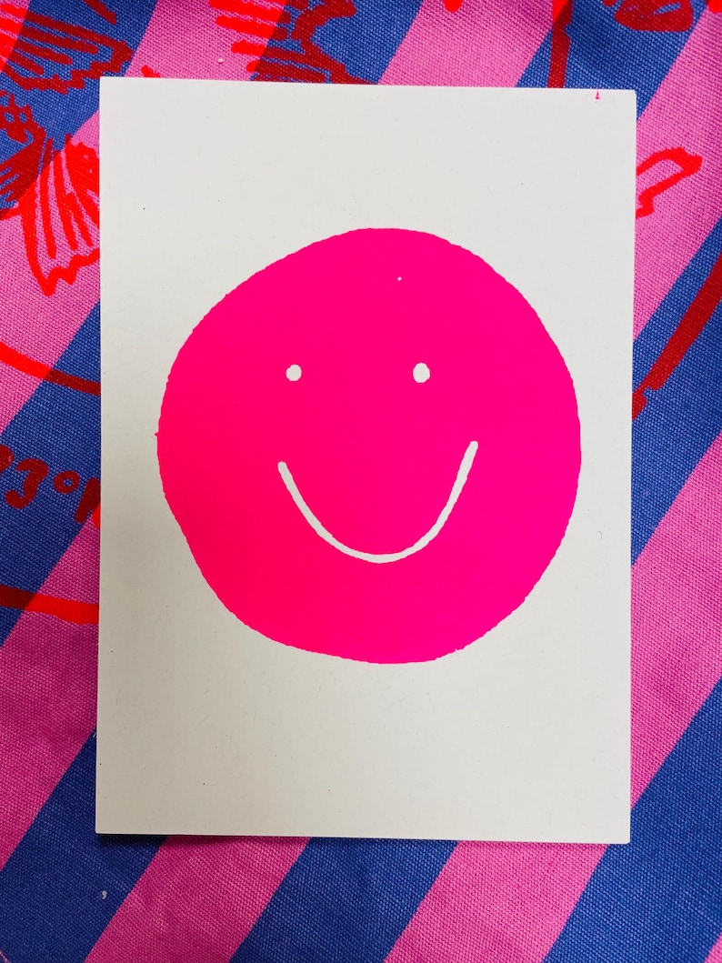 K&ouml;nnte beinhalten: Eine wei&szlig;e rechteckige Karte mit einem gro&szlig;en, leuchtend pinkfarbenen Smiley-Gesicht. Das Smiley-Gesicht hat zwei kleine wei&szlig;e Punkte f&uuml;r die Augen und eine gebogene wei&szlig;e Linie f&uuml;r den Mund. Die Karte befindet sich vor einem gemusterten Hintergrund.