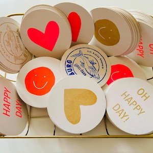 May include: A collection of round coasters with various designs, including a neon pink heart, a gold heart, a smiley face, and the words "OH HAPPY DAY!". Some coasters feature a fish illustration and the text "keep cool!". The coasters are in a gold wire basket.