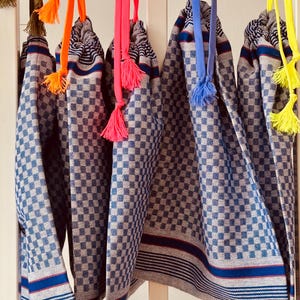 Puede incluir: Varias toallas con estampado de cuadros azules y blancos con coloridos detalles de borlas cuelgan de una superficie blanca. Las toallas tienen rayas azules y blancas en el borde inferior. Las borlas son de color naranja, rosa, azul y amarillo.