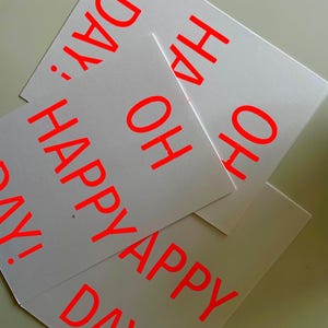 K&ouml;nnte beinhalten: Mehrere wei&szlig;e Karten mit dem Aufdruck "OH HAPPY DAY!" in leuchtendem Rot. Die Karten &uuml;berlappen sich leicht, wodurch ein geschichteter Effekt entsteht. Der Text ist fett und gut lesbar, was ein fr&ouml;hliches Design ergibt.