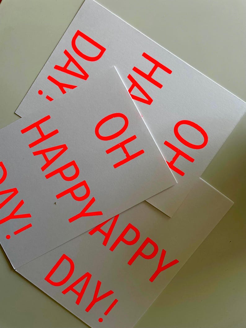 K&ouml;nnte beinhalten: Mehrere wei&szlig;e Karten mit dem Aufdruck "OH HAPPY DAY!" in leuchtendem Rot. Die Karten &uuml;berlappen sich leicht, wodurch ein geschichteter Effekt entsteht. Der Text ist fett und gut lesbar, was ein fr&ouml;hliches Design ergibt.