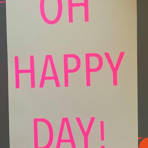 K&ouml;nnte beinhalten: Ein rechteckiges Schild mit den Worten "OH HAPPY DAY!" in leuchtend pinkfarbenen Buchstaben. Der Text ist auf einem hellgrauen Hintergrund zentriert, mit einem dunkelgrauen Rand und orangefarbenen Akzenten.