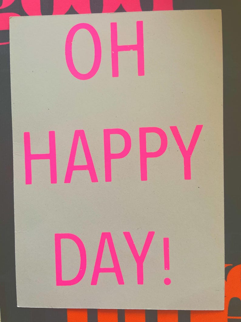 K&ouml;nnte beinhalten: Ein rechteckiges Schild mit den Worten "OH HAPPY DAY!" in leuchtend pinkfarbenen Buchstaben. Der Text ist auf einem hellgrauen Hintergrund zentriert, mit einem dunkelgrauen Rand und orangefarbenen Akzenten.