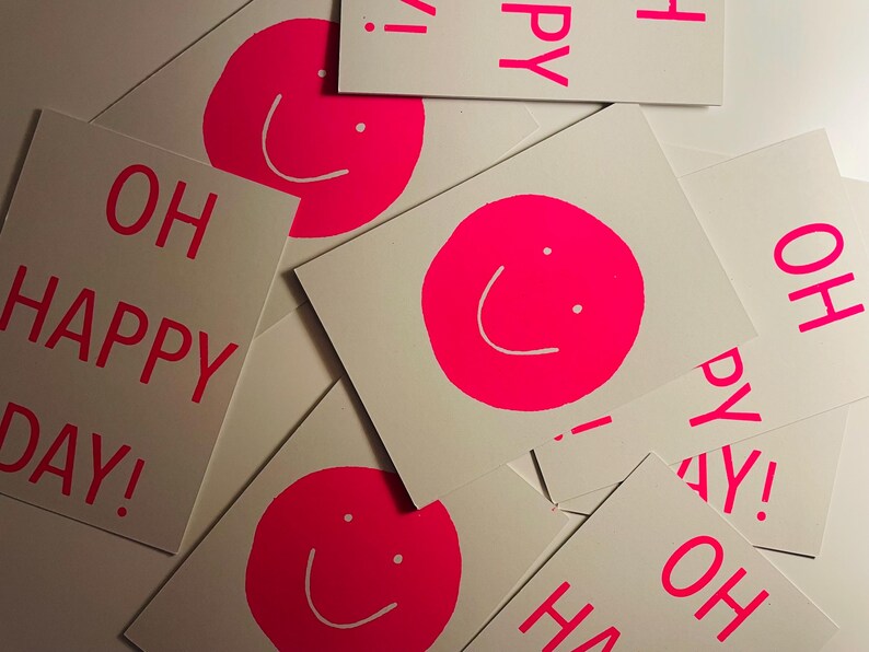 K&ouml;nnte beinhalten: Eine Ansammlung wei&szlig;er Karten mit einem leuchtend pinkfarbenen Smiley-Gesicht und dem pinkfarbenen Schriftzug "OH HAPPY DAY!". Die Karten sind verstreut, was eine fr&ouml;hliche und verspielte Anordnung ergibt.