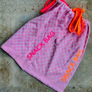 May include: Two pink snack bags with a checkered pattern and the words "SNACK BAG" printed in neon orange and pink. The bags have drawstring closures with neon orange and pink cords.