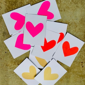 K&ouml;nnte beinhalten: Eine Ansammlung wei&szlig;er Karten mit herzf&ouml;rmigen Designs in leuchtendem Pink, Rot und Gelb. Die Karten liegen auf einer strukturierten, goldbraunen Oberfl&auml;che und bilden ein lebendiges Arrangement.