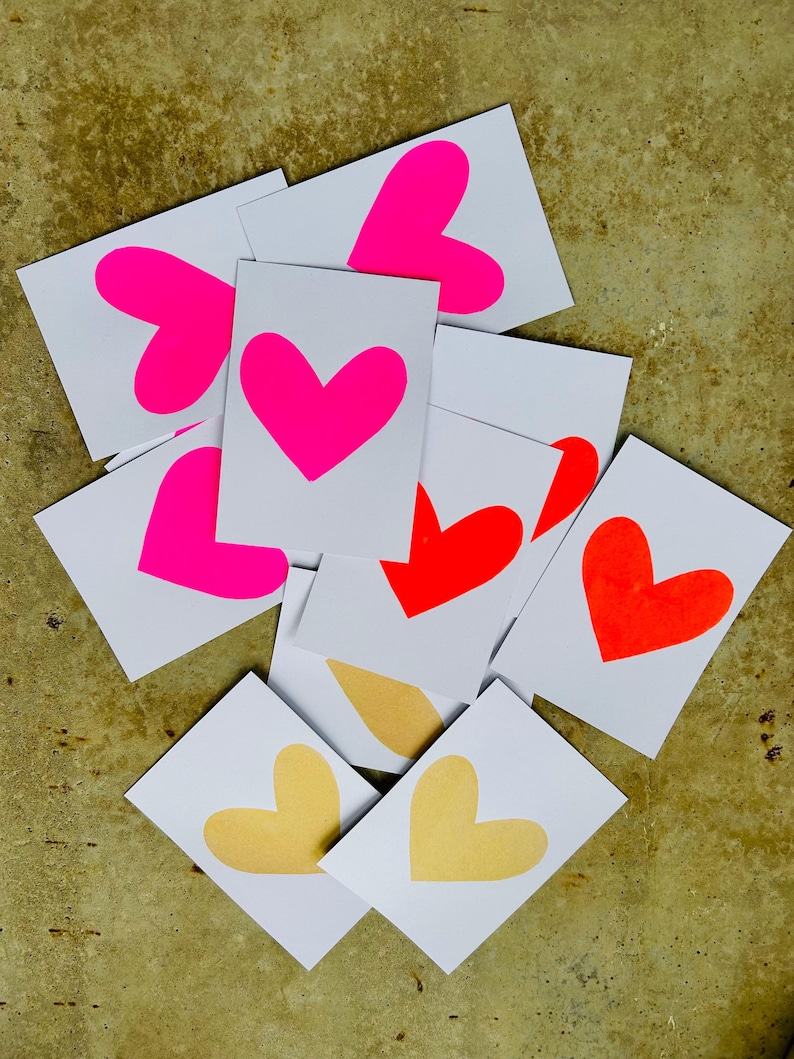 K&ouml;nnte beinhalten: Eine Ansammlung wei&szlig;er Karten mit herzf&ouml;rmigen Designs in leuchtendem Pink, Rot und Gelb. Die Karten liegen auf einer strukturierten, goldbraunen Oberfl&auml;che und bilden ein lebendiges Arrangement.