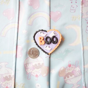 K&ouml;nnte beinhalten: Eine lila und orangefarbene herzf&ouml;rmige Anstecknadel mit dem Wort "BOO" in schwarzen Buchstaben und wei&szlig;en Geister-Silhouetten. Die Anstecknadel befindet sich auf einem hellblauen Stoff mit einem pastellfarbenen Regenbogen-, Herz- und Sternmuster.
