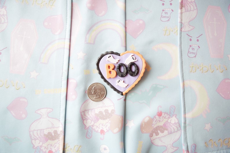 K&ouml;nnte beinhalten: Eine lila und orangefarbene herzf&ouml;rmige Anstecknadel mit dem Wort "BOO" in schwarzen Buchstaben und wei&szlig;en Geister-Silhouetten. Die Anstecknadel befindet sich auf einem hellblauen Stoff mit einem pastellfarbenen Regenbogen-, Herz- und Sternmuster.