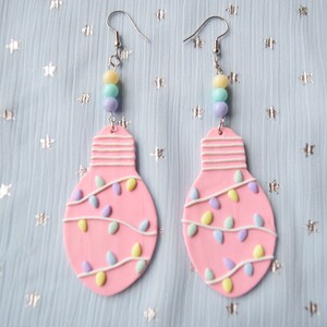May include: Pink lightbulb shaped earrings with white and pastel colored string lights and beads hanging from the bottom.