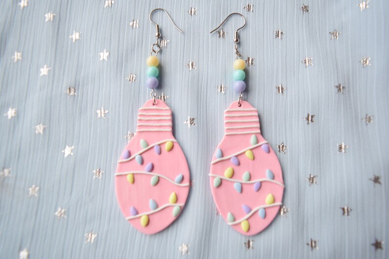 May include: Pink lightbulb shaped earrings with white and pastel colored string lights and beads hanging from the bottom.