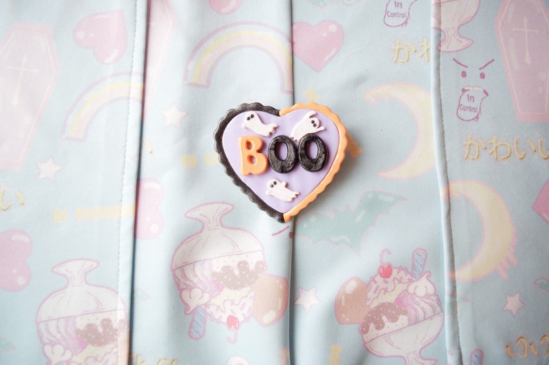 K&ouml;nnte beinhalten: Eine herzf&ouml;rmige Anstecknadel in Lila und Schwarz mit dem Wort "BOO" in orangefarbenen und schwarzen Buchstaben. Auf der Anstecknadel befinden sich kleine wei&szlig;e Geisterfiguren.