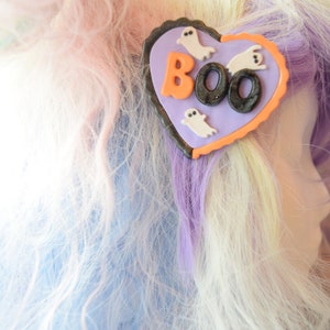 K&ouml;nnte beinhalten: Eine lila und orangefarbene herzf&ouml;rmige Haarklammer mit dem Wort "BOO" in schwarzen und orangen Buchstaben und drei wei&szlig;en Geister-Silhouetten.