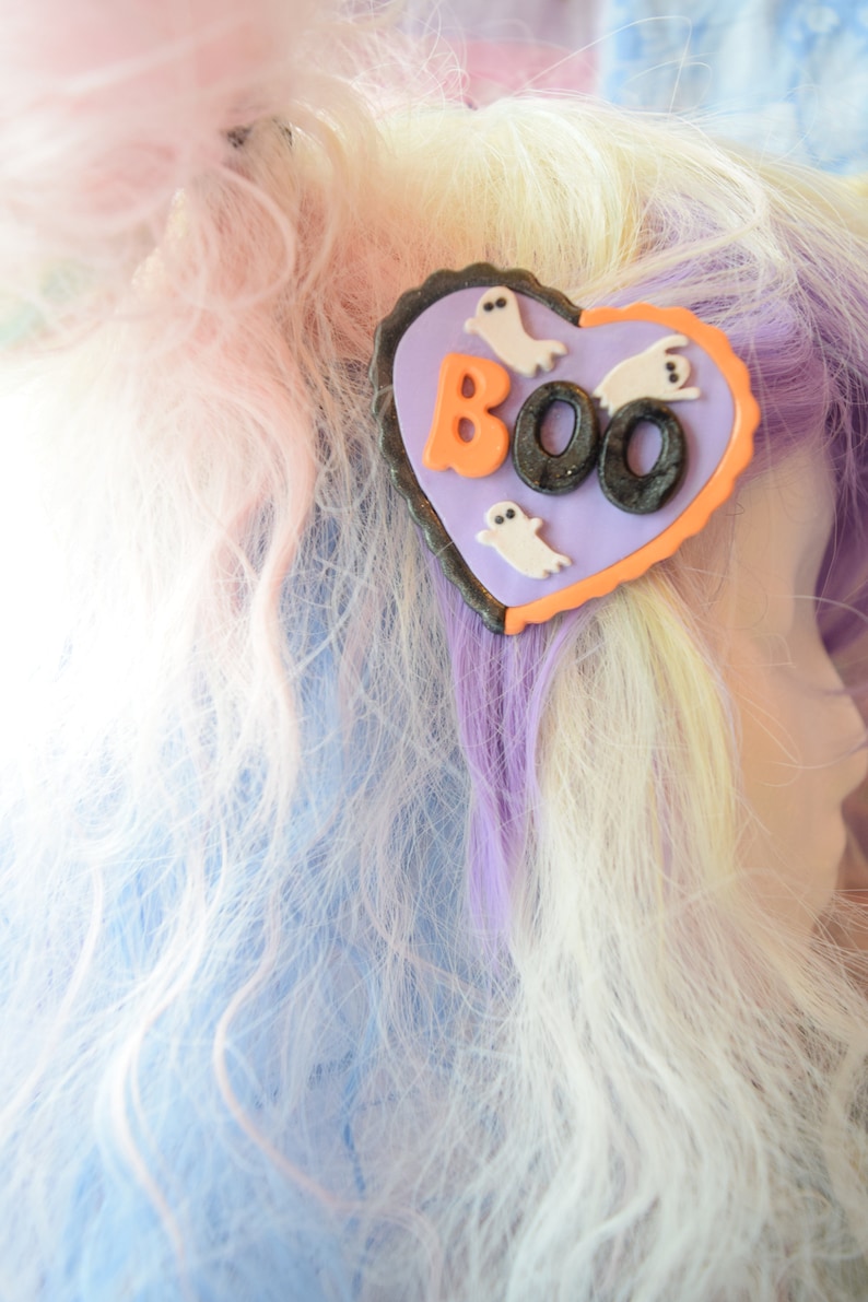 K&ouml;nnte beinhalten: Eine lila und orangefarbene herzf&ouml;rmige Haarklammer mit dem Wort "BOO" in schwarzen und orangen Buchstaben und drei wei&szlig;en Geister-Silhouetten.