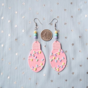 May include: A pair of pink lightbulb earrings with pastel colored string lights and silver hooks. The earrings are shown next to a US quarter for size reference.