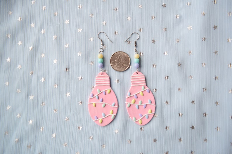 May include: A pair of pink lightbulb earrings with pastel colored string lights and silver hooks. The earrings are shown next to a US quarter for size reference.