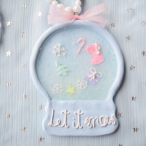 May include: A blue and white snow globe ornament with a pink ribbon and the words "Let it snow" on the bottom. The snow globe contains a small Christmas tree, a candy cane, a stocking, and snowflakes.
