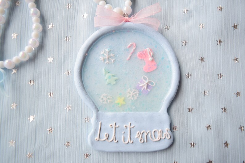 May include: A blue and white snow globe ornament with a pink ribbon and the words "Let it snow" on the bottom. The snow globe contains a small Christmas tree, a candy cane, a stocking, and snowflakes.
