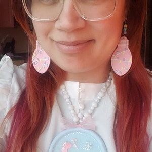 May include: A woman wearing a pink beret, clear glasses, and a white blouse with a pearl necklace. She is wearing pink lightbulb earrings and a blue and white snow globe charm necklace with the text "Let it snow".