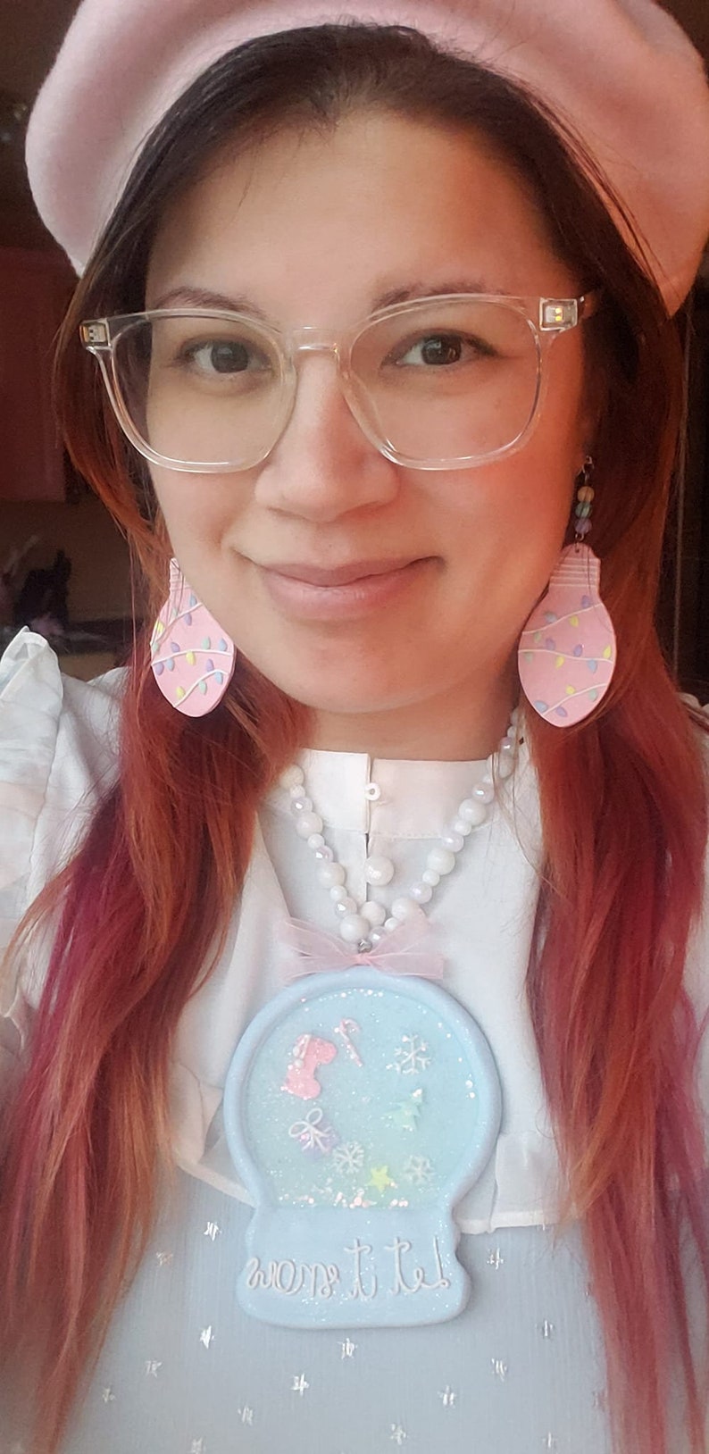 May include: A woman wearing a pink beret, clear glasses, and a white blouse with a pearl necklace. She is wearing pink lightbulb earrings and a blue and white snow globe charm necklace with the text "Let it snow".