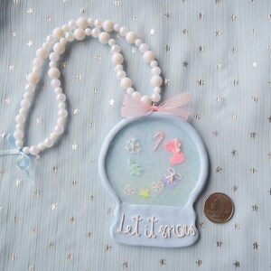 May include: A blue and white snow globe necklace with a pink bow and the text "Let it snow". The necklace is made of white beads and has a blue background with white snowflakes and a pink candy cane.