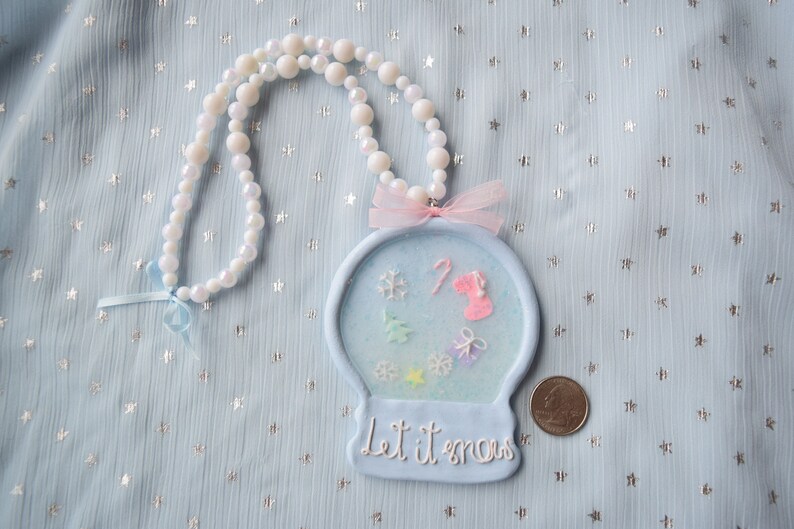May include: A blue and white snow globe necklace with a pink bow and the text "Let it snow". The necklace is made of white beads and has a blue background with white snowflakes and a pink candy cane.