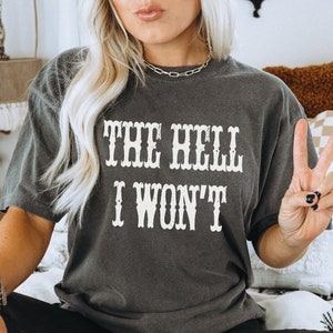 May include: A woman wearing a dark gray t-shirt with the text "THE HELL I WON'T" in white. She is wearing black leather pants and a brown beanie.