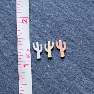 May include: Three small metal cactus charms in silver, gold, and copper tones. The charms are arranged in a row on a dark gray surface with a measuring tape in the background.