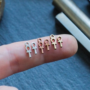 May include: A close-up of a finger holding a row of seven small, silver, gold, and rose gold Ankh charms. The charms are arranged in a line from left to right, with the silver charms on the left and the gold charms on the right.