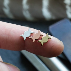 May include: Three crescent moon earrings in silver, rose gold, and gold. The earrings are shown on a finger.