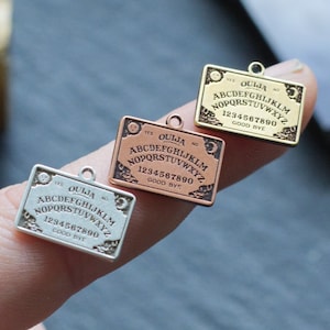 May include: Three miniature Ouija board charms in silver, rose gold, and gold. Each charm features the letters of the alphabet, numbers, and the words "Ouija", "Yes", "No", and "Goodbye".